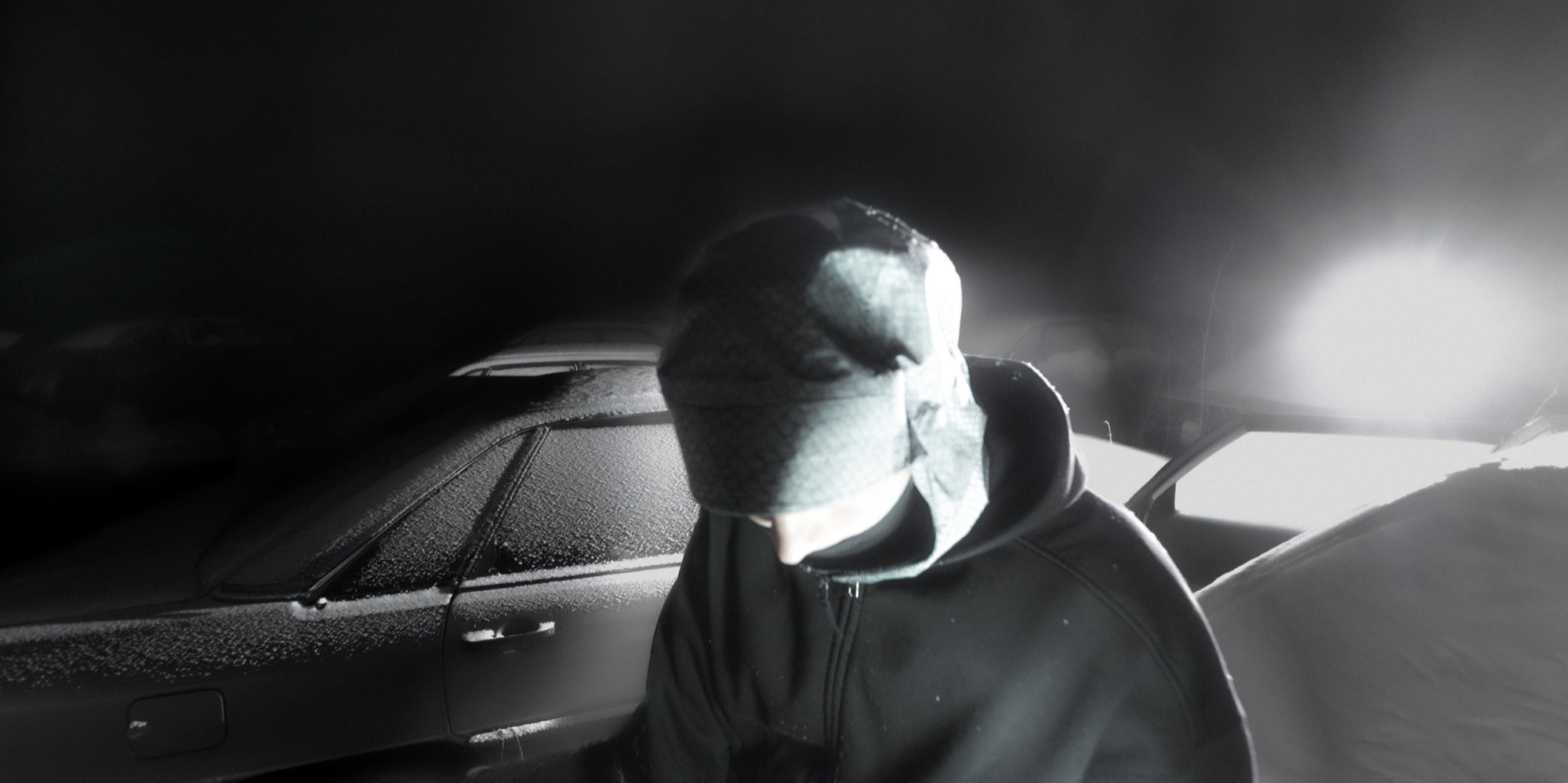 Masked figure in front of a car and bright light.
